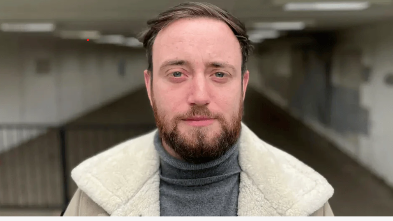 Jacob tem barba e está usando um suéter cina e uma jaqueta bege por cima