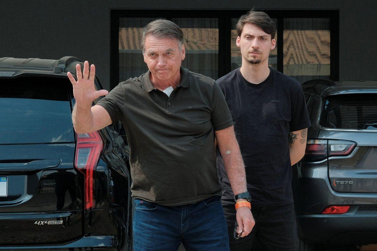 Jair Bolsonaro em pé, acenando, entre carros; atrás dele, Jair Renan