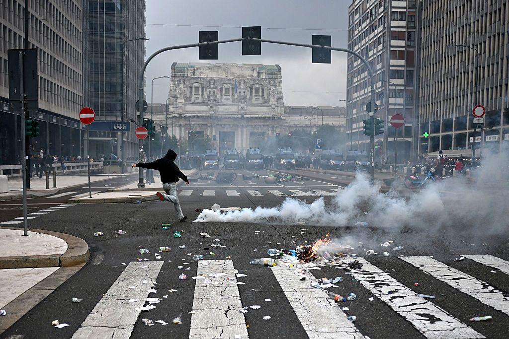 Un manifestante de espaldas hace el gesto de dar una patada en medio de una calle de Milán, con humo a su derecha y vehículos de la policía antidisturbios al fondo, cerca de la estación de tren Milano Centrale.