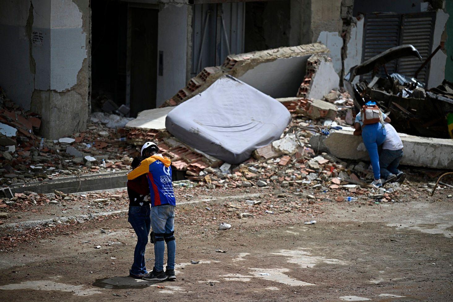 Un hombre con una chaqueta con los colores de la bandera de Venezuela abraza a una mujer. A unos cuantos metros, una mujer abraza a un hombre que está de rodillas. Todos están entre los escombros frente a un edificio en ruinas. Hay un colchón

