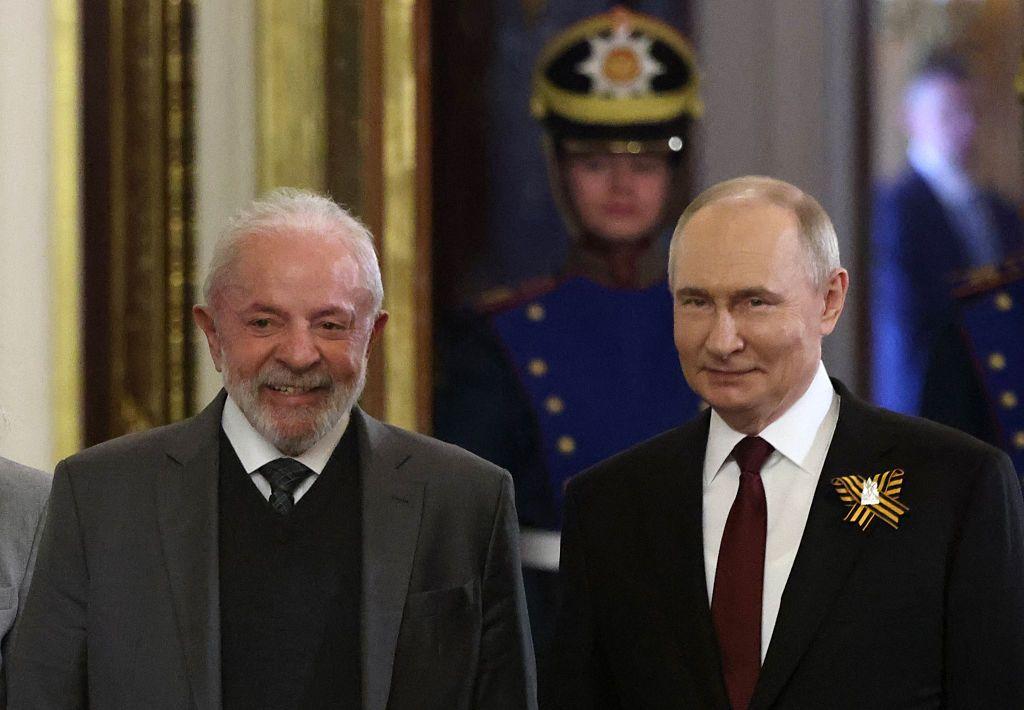 Lula sonriente camina junto a Vladimir Putin. Lula lleva una chaqueta de traje gris oscuro, con un chaleco oscuro, una camisa blanca y corbata. Putin luca una chaqueta oscuro en cuya solapa tiene un prendedor con rayas amarillas, junto a una camisa blanca y una corbata vino tinto.