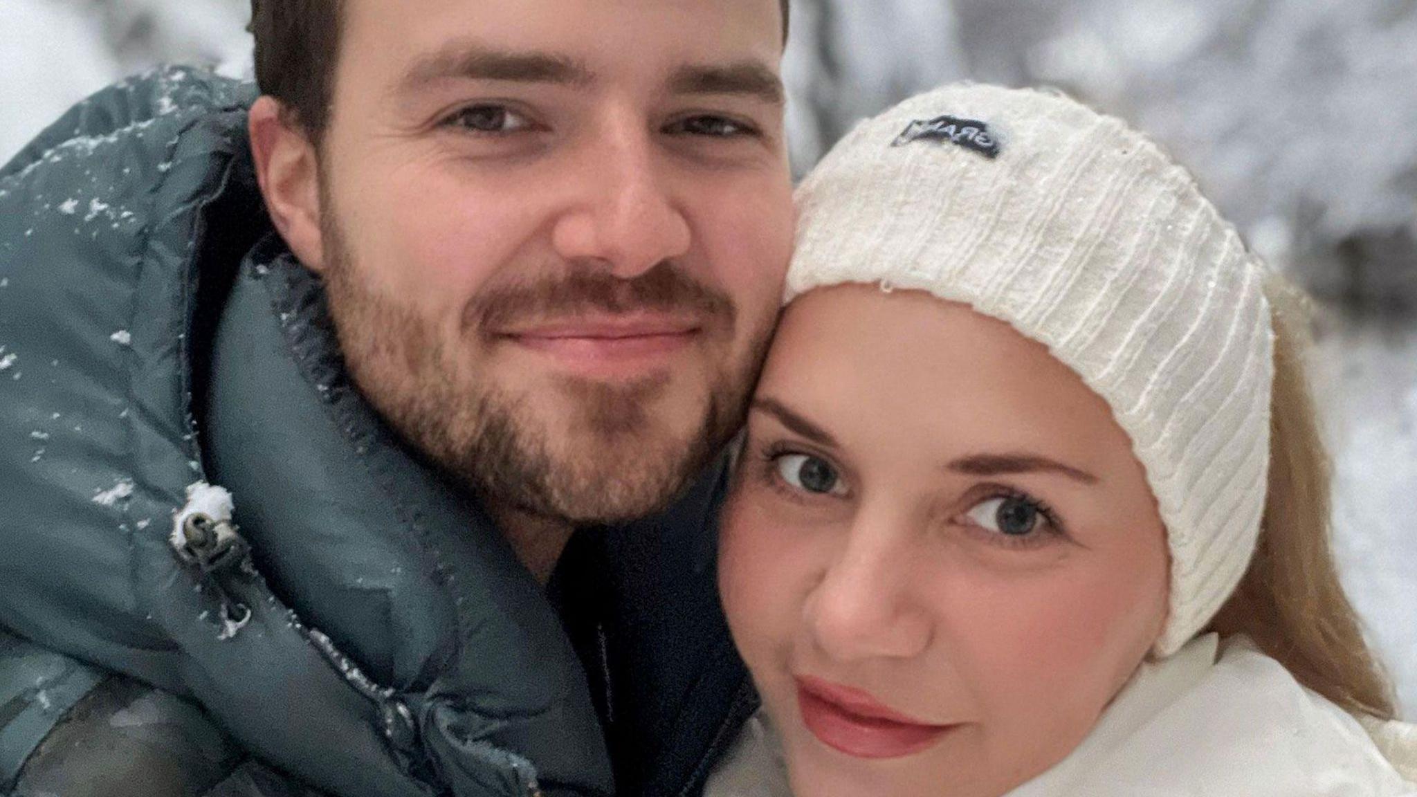 Una pareja heterosexual abrazada y sonriendo levemente para la cámara mientras lucen atuendos de invierno y se vislumbra un paisaje nevado en el fondo.