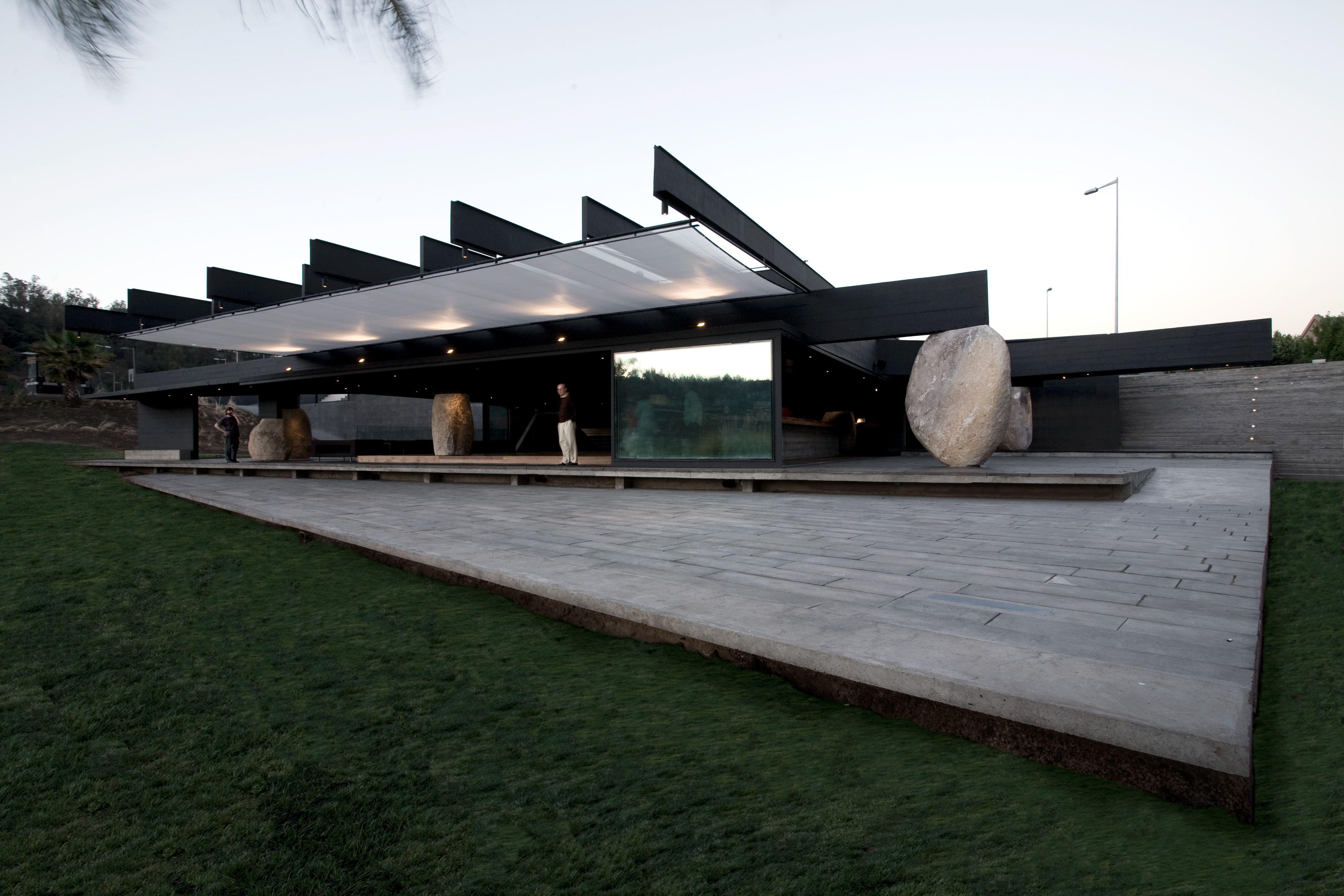 Edificio de líneas modernas con techo en voladizo, grandes bloques de piedra y amplias superficies de vidrio sobre una plataforma inclinada.