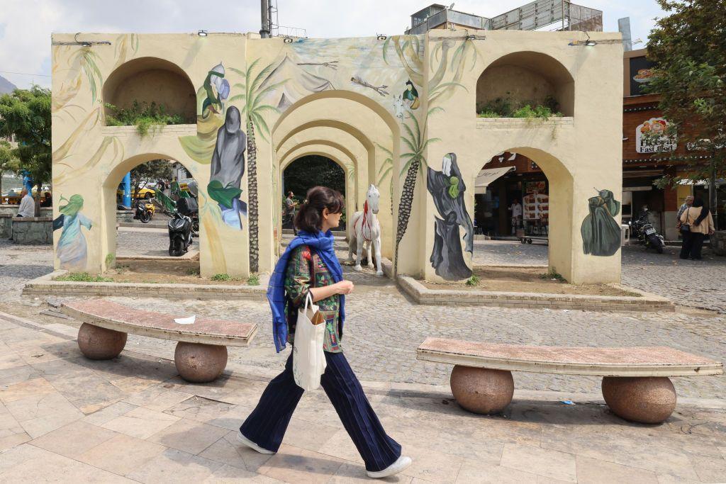 امرأة تنظر بعيداً عن الكاميرا، وتسير في أحد شوارع طهران مرتديةً الجينز وقميصاً مُلوناً بدون غطاء على رأسها، وتحمل حقيبة بيضاء . 