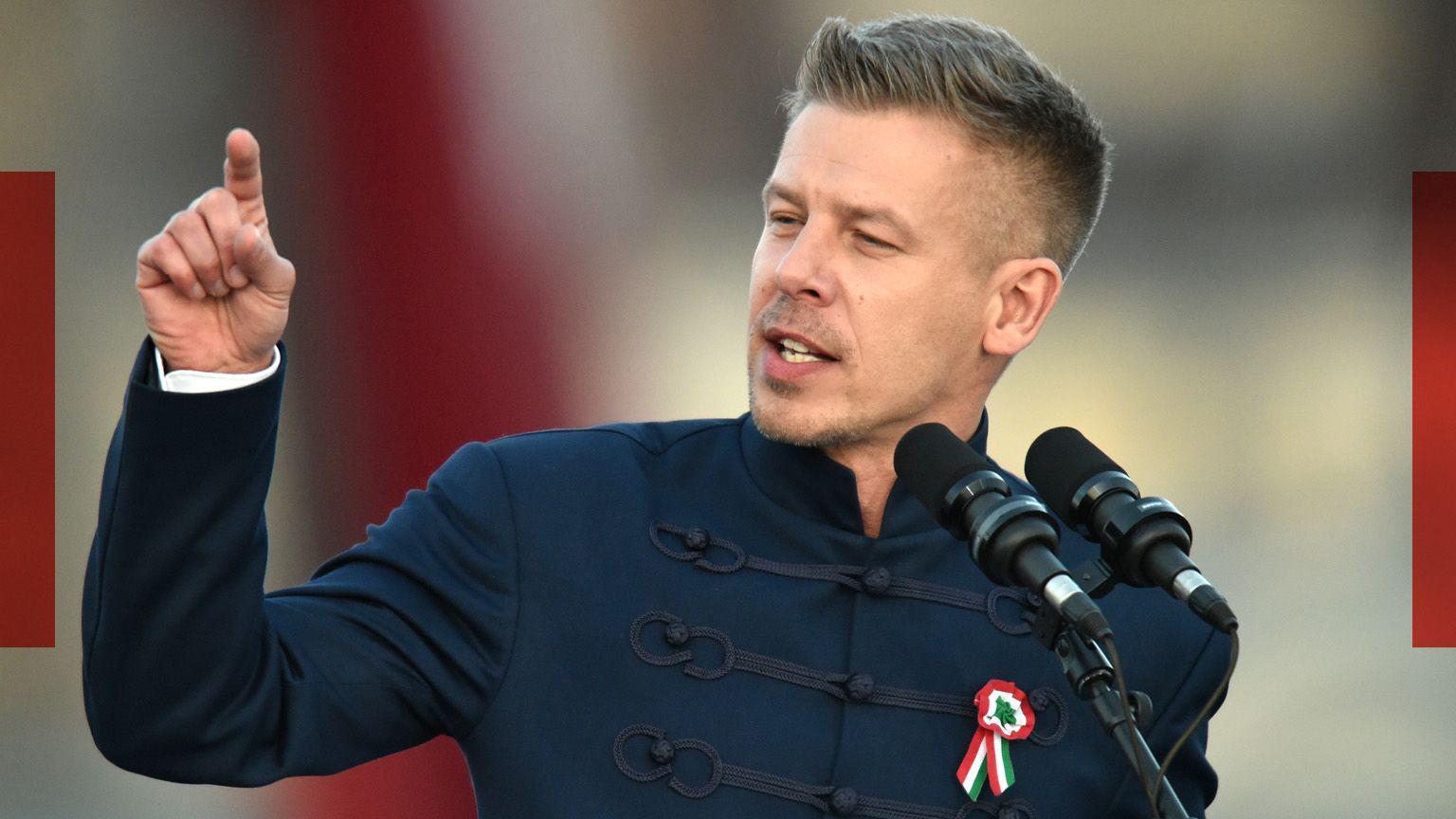 Peter Magyar gesticula em um p&oacute;dio. Ele veste um palet&oacute; azul de gola alta com brocado na frente e tem uma roseta presa ao peito.