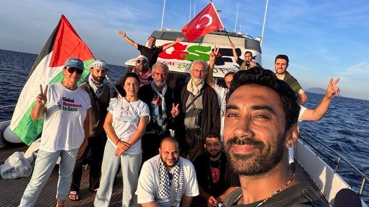 Nasmejani čovek pravi selfi na brodu, dok u pozadini pozira još desetak ljudi. Vide se turska i palestinska zastava, kao i transparent GSF-a u bojama palestinske zastave.
