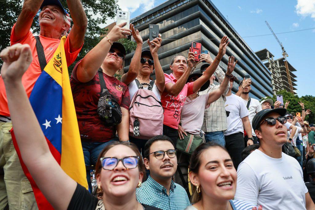 Una multitud de personas, una de ellas con una bandera de Venezuela, sonríen. Varios levantan sus teléfonos celulares como si estuvieran grabando un video. Están en una calle al aire libre y al fondo se ve un edificio alto..