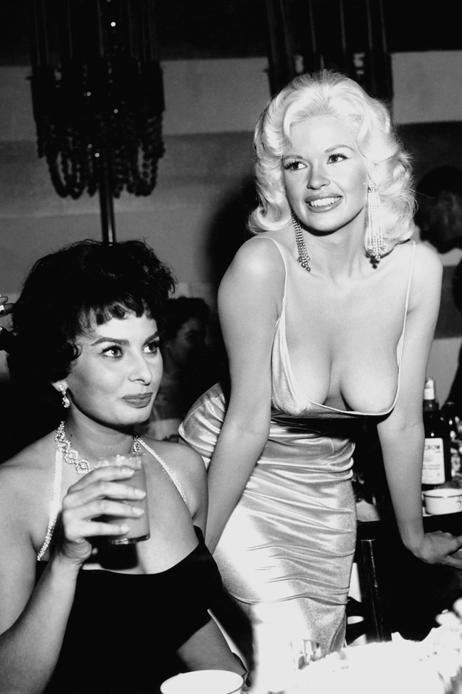 Sofía Loren, con un vestido negro con un amplio escote, está sentada y tiene un vaso en la mano. A su lado, parada y sonriendo, está Jayne Mansfield, que lleva un vestido de seda blanco con un amplio escote