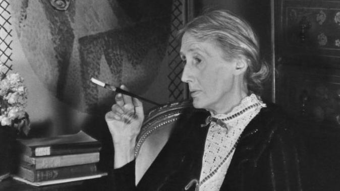 Retrato de la escritora británica Virginia Woolf (1882-1941) fumando con una boquilla y un libro abierto sobre su regazo, Londres, Inglaterra, 1939. En la pared, detrás de ella, hay un fresco de su hermana, Vanessa Bell.