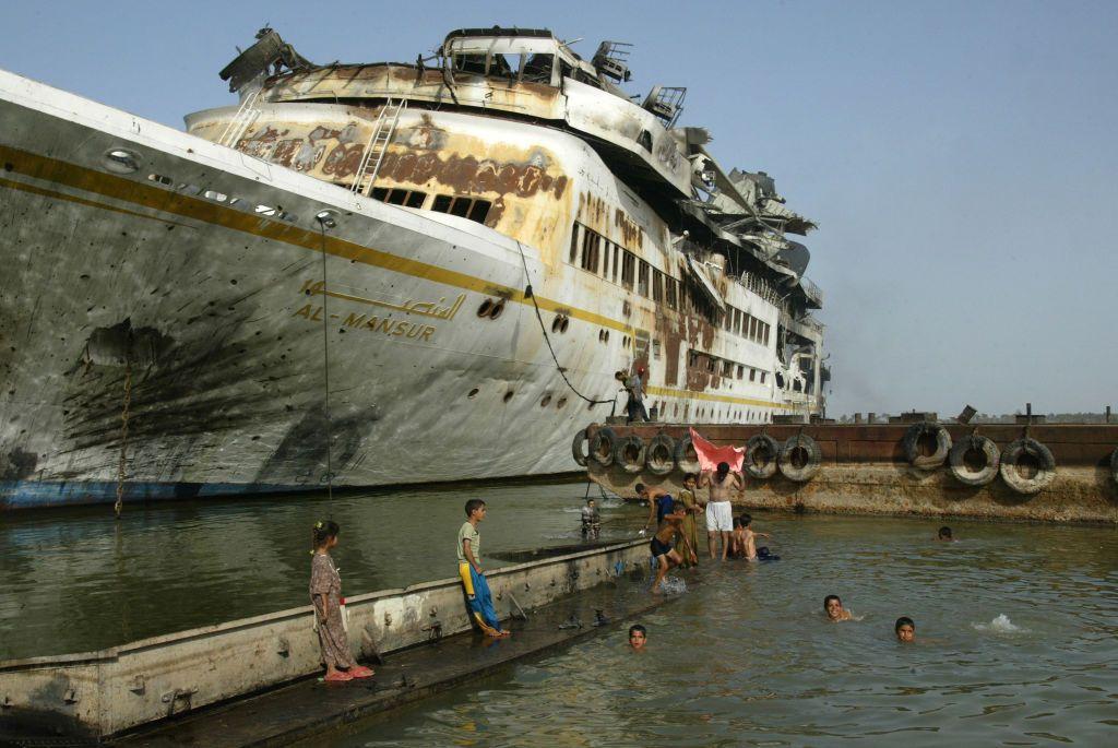 Un grupo de adultos y niños se bañan en el puerto de Basora junto a un barco abandonado y oxidado. 