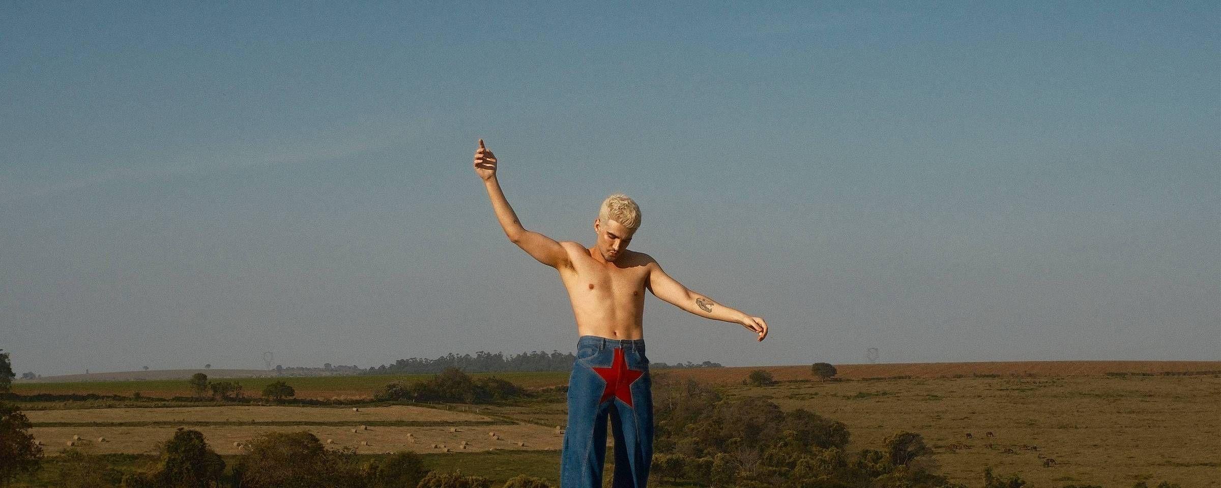 Jão, um homem sem camisa, de cabelo platinado, está em pé em uma paisagem rural, com os braços erguidos e a cabeça inclinada para baixo. Ele veste uma calça jeans com uma grande estrela vermelha na parte frontal, enquanto ao fundo aparecem campos, árvores e um céu aberto.