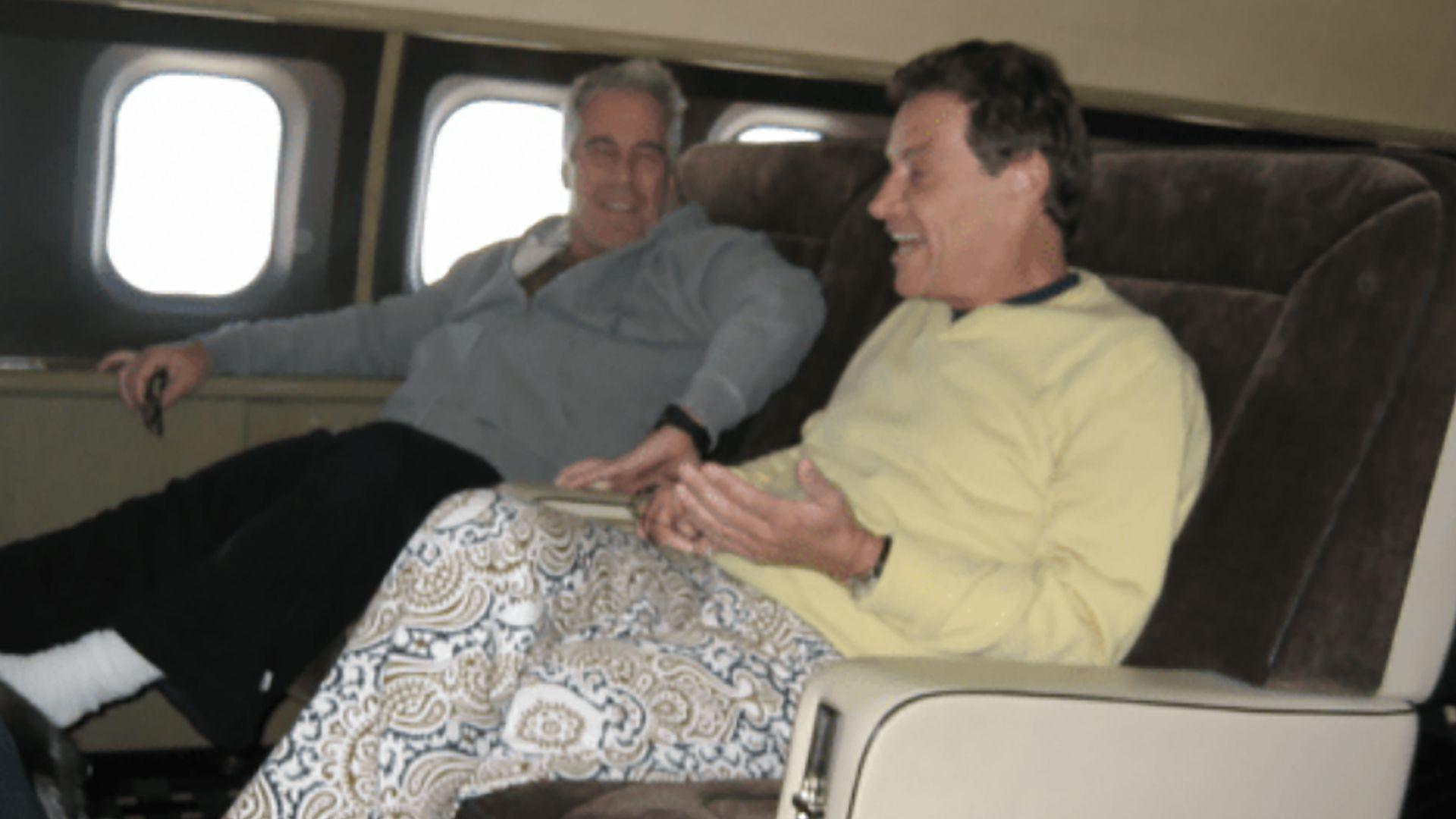 兩名面帶微笑的男子坐在飛機上舒適的大座椅上。愛潑斯坦留著灰白短髮，穿著灰色上衣和黑色長褲。布魯內爾留著深色短髮，身穿淺黃色上衣和長褲，褲子以白色為底，飾有深藍色和金色圖案。