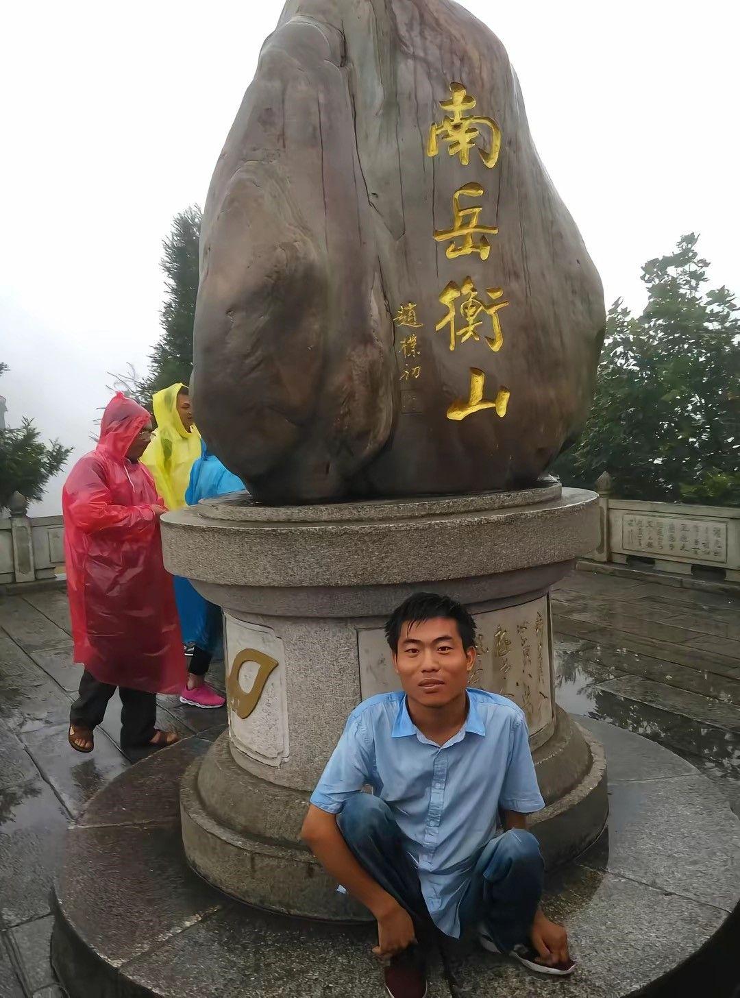 El doctor Li, bajo la lluvia, en un punto de la ruta del monte Henshang. Lleva una camisa azul y unos jeans, detrás de él hay una piedra gigante que se levanta como en una especie de pedestal