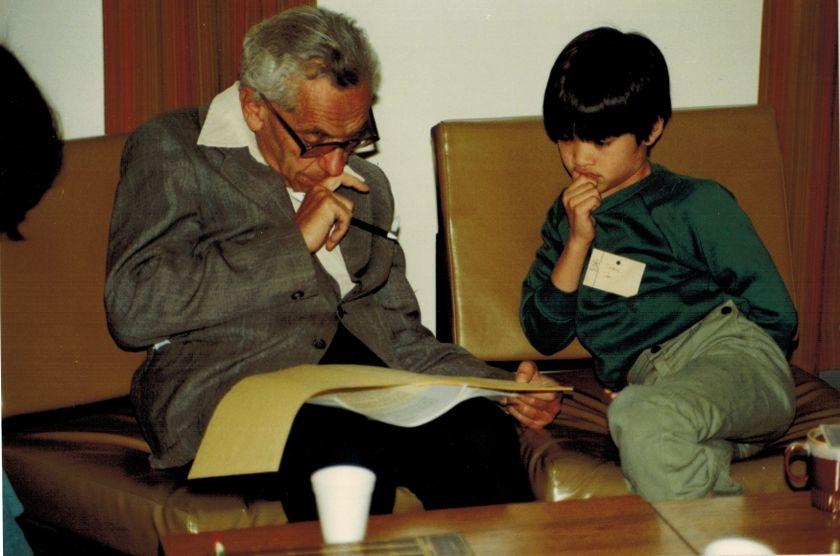 Paul Erdős, con lentes, está sentado leyendo unos papeles que tiene sobre sus piernas, en una mano tiene un bolígrafo. A su lado, Terence Tao también observa los papeles
