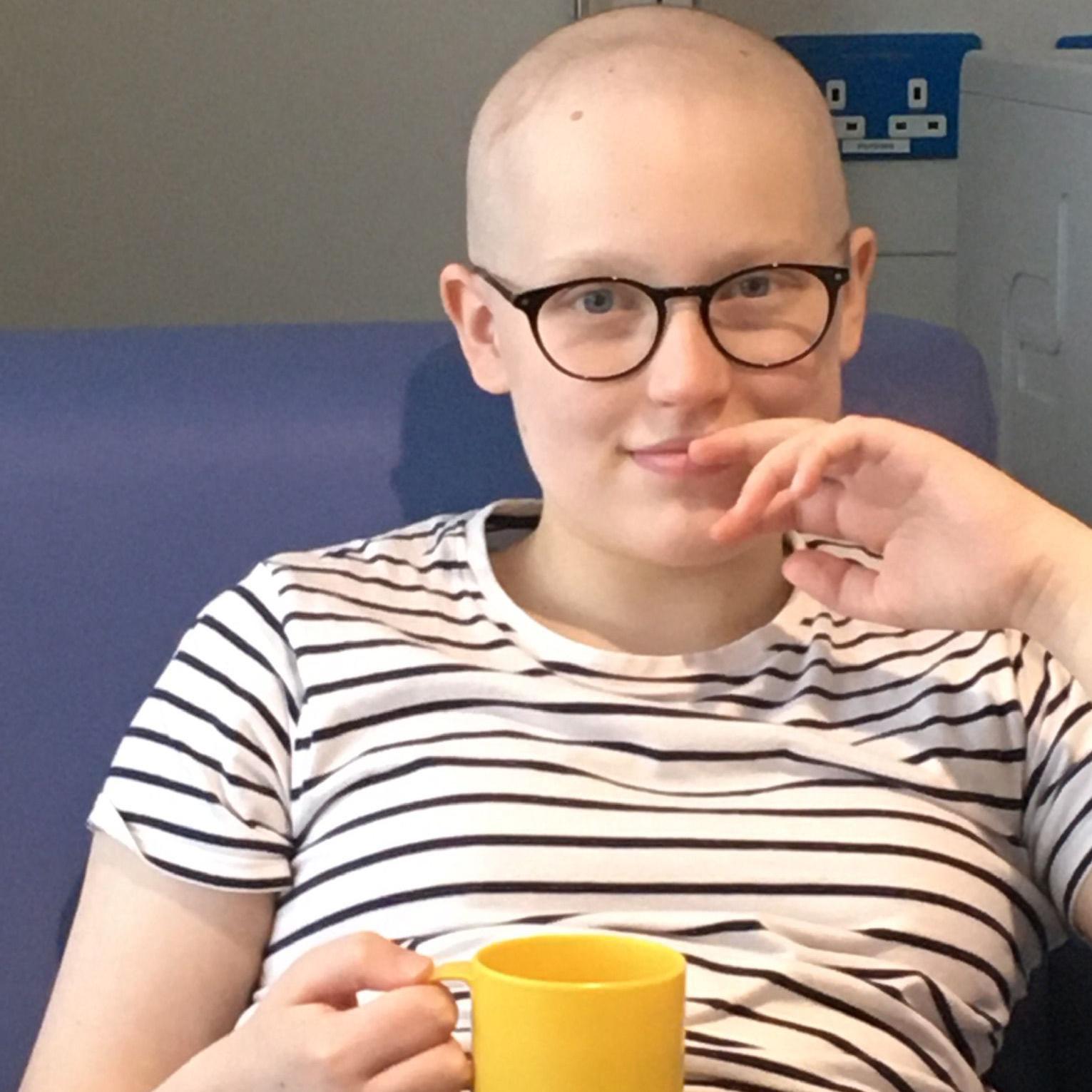 Una joven con gafas y una camiseta de rayas blancas y negras sostiene una taza amarilla y está sentada en un sofá azul. No tiene pelo.