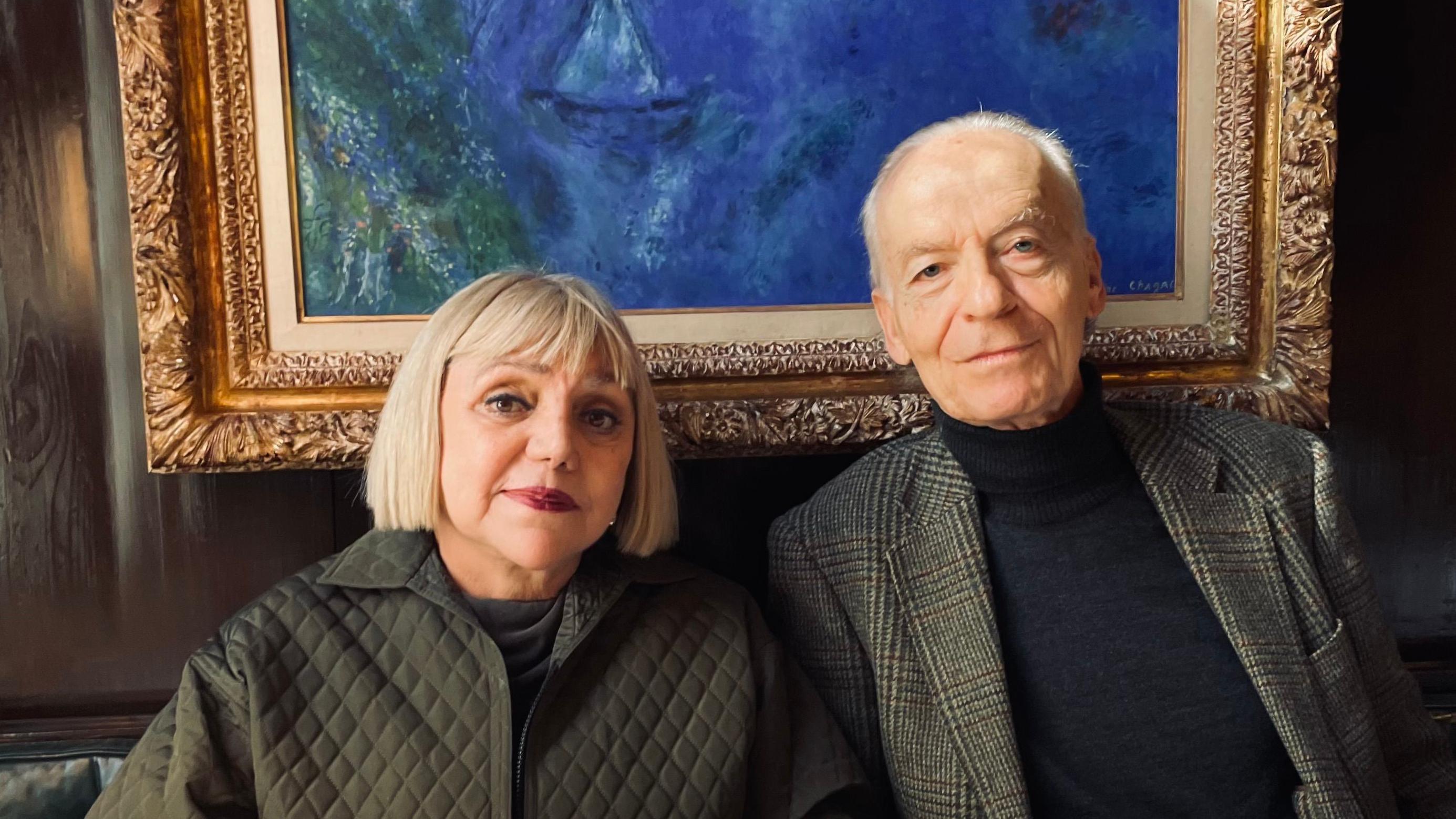 Una mujer madura con pelo claro y un hombre maduro de pelo canoso vestidos causual pero elegantemente y sentados frente a un  cuadro  de Chagall