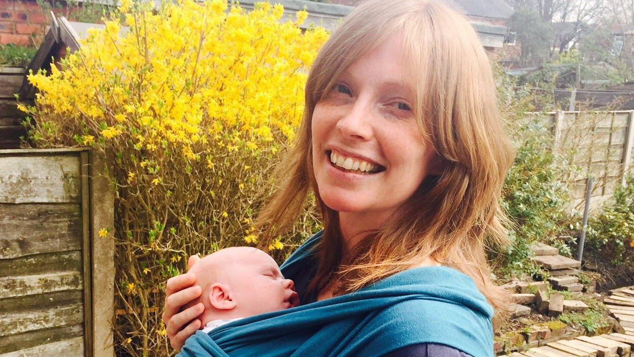 Helen Ledwick sorri para a câmera com um bebê em um sling.