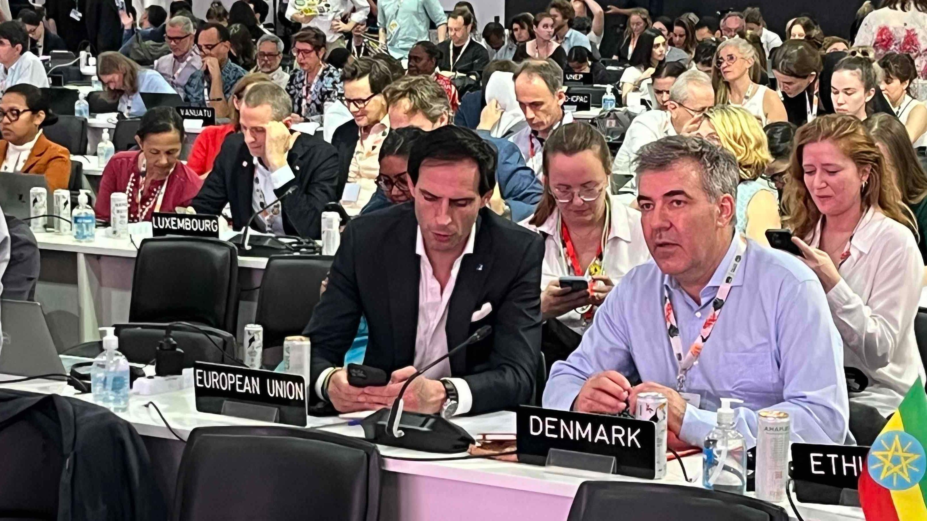 Miembros del equipo negociador de la Unión Europea durante el plenario. Uno mira hacia el podio, otro consulta su teléfono.