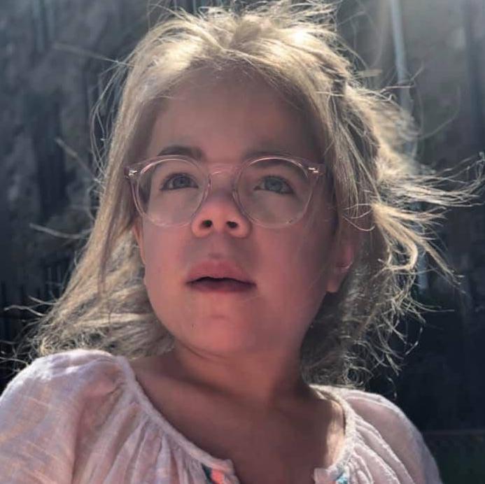 Sophia mira ligeramente fuera de cámara. Tiene el cabello largo y rubio recogido hacia atrás, y lleva unas gafas grandes de montura rosa y una blusa blanca de verano.