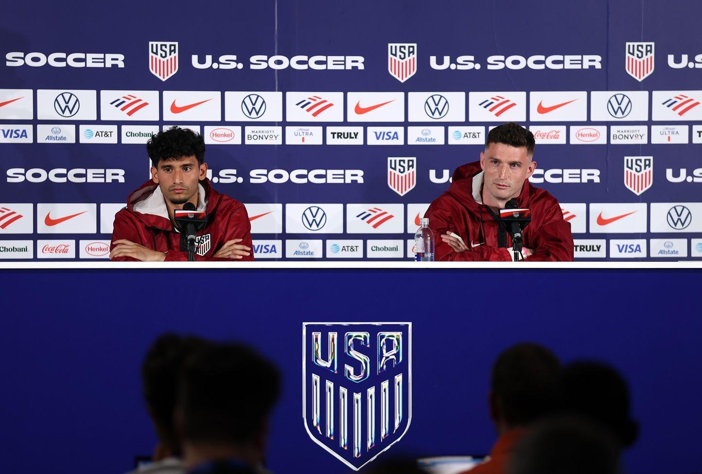 Ricardo Pepi y Matthew Freese con chaquetas rojas del uniforme de la selección de EE.UU. Ambos están sentados y tienen micrófonos al frente. Detrás tienen una pared llena de símbolos de marcas y escudos de USA y las palabras U.S. Soccer. Al frente hay periodistas

