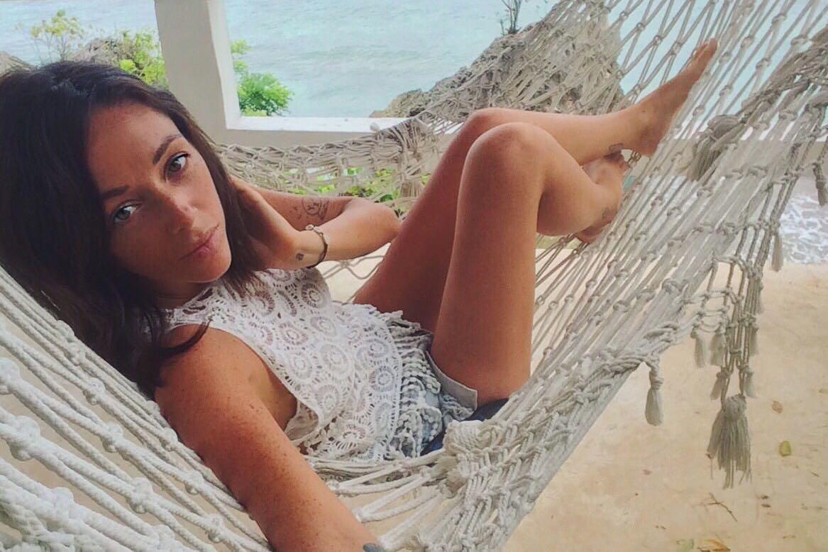 Kirsty McKie está sentada en una hamaca y mira a la cámara mientras se hace un selfi. Al fondo se ve el mar y un árbol, y debajo de ella hay arena. Tiene el pelo castaño que le llega hasta los hombros.