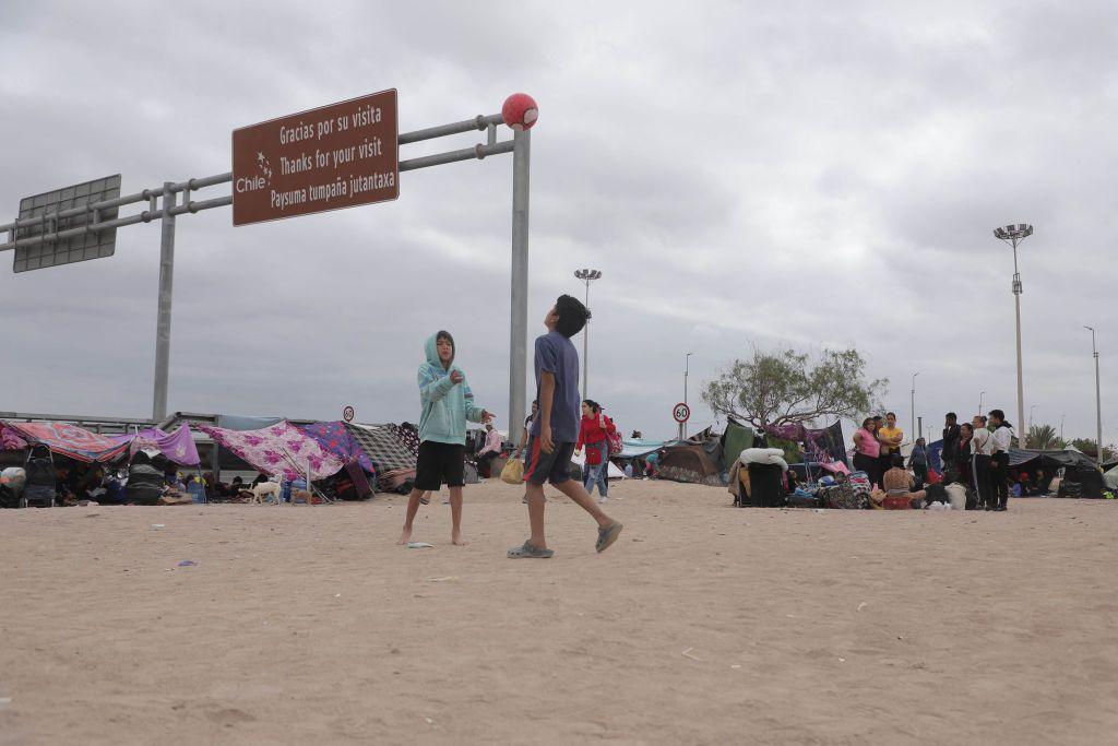 Niños jugando con una pelota en un área de arena junto a carpas, con un letrero fronterizo de Chile al fondo.