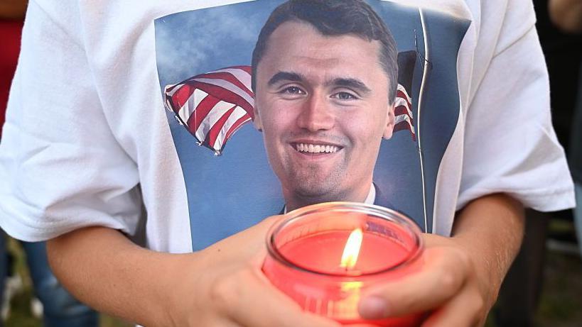 Los residentes de Lemont, Illinois, se reúnen en un parque sereno para rendir homenaje a Charlie Kirk durante una vigilia el 11 de septiembre de 2025. En la imagen se ve en primer plano unas manos que sostienen una vela encendida. La persona que la sostiene lleva una camiseta con una foto estampada con la cara de Charlie Kirk. 