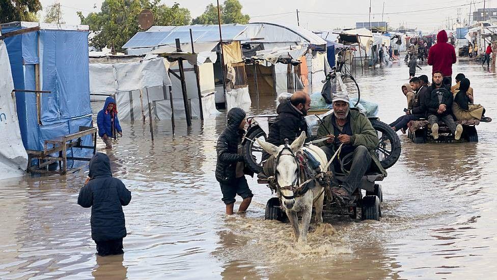 Una camino inundado entre tiendas de campaña. Se ve a un hombre en un carro tirado por un caballo flaco intentando avanzar en el agua