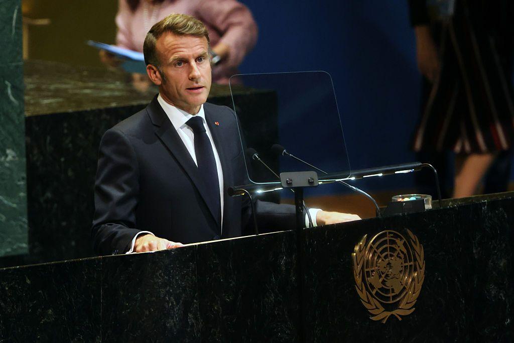 يرتدي ماكرون بدلة رسمية سوداء وقميصاً أبيض وربطة عنق سوداء، يقف على منصة الأمم المتحدة متحدثاً.