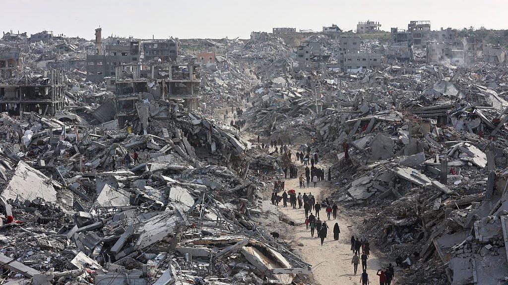 A line of people walk down a dirt road, surrounded on each side by tall piles of destroyed buildings. 