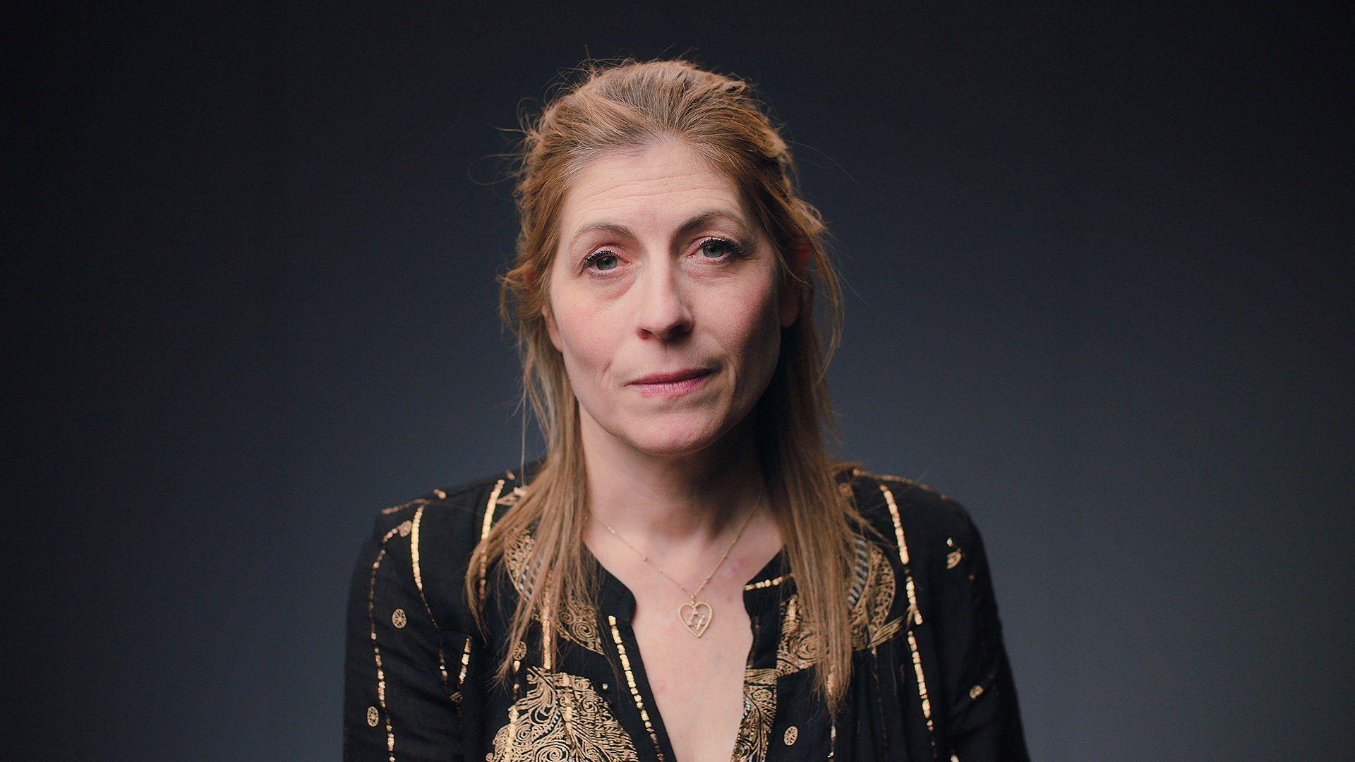 امرأة ذات شعر طويل بني فاتح مائل للحمرة تنظر إلى الأمام مباشرة. ترتدي بلوزة سوداء بخيوط ذهبية. وتضع حول عنقها نجمة داود داخل قلب.