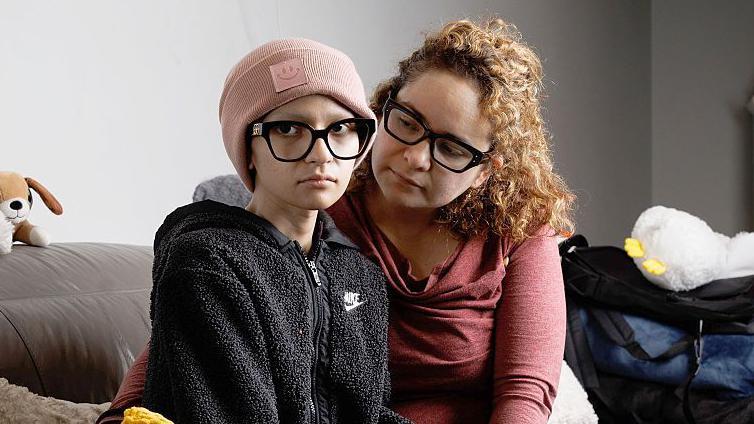 Ofelia, delgada y luciendo un gorro, mira a la cámara con una expresión de tristeza mientras su madre la mira y la rodea con sus brazos.