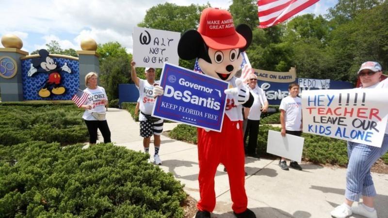 Persona disfrazada de mikey mouse con un letrero que dice DeSantis. Otras personas con carteles a su alrededor realizan una protesta contra disney
