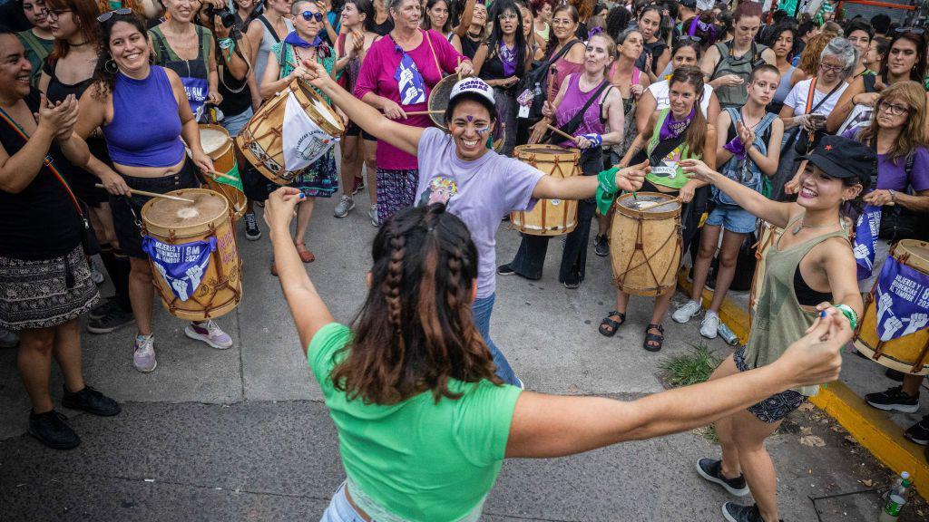 Três mulheres dançando de mãos dadas, no que parece ser uma manifestação feminista. Ao fundo, outras mulheres olham para elas e tocam tambores.