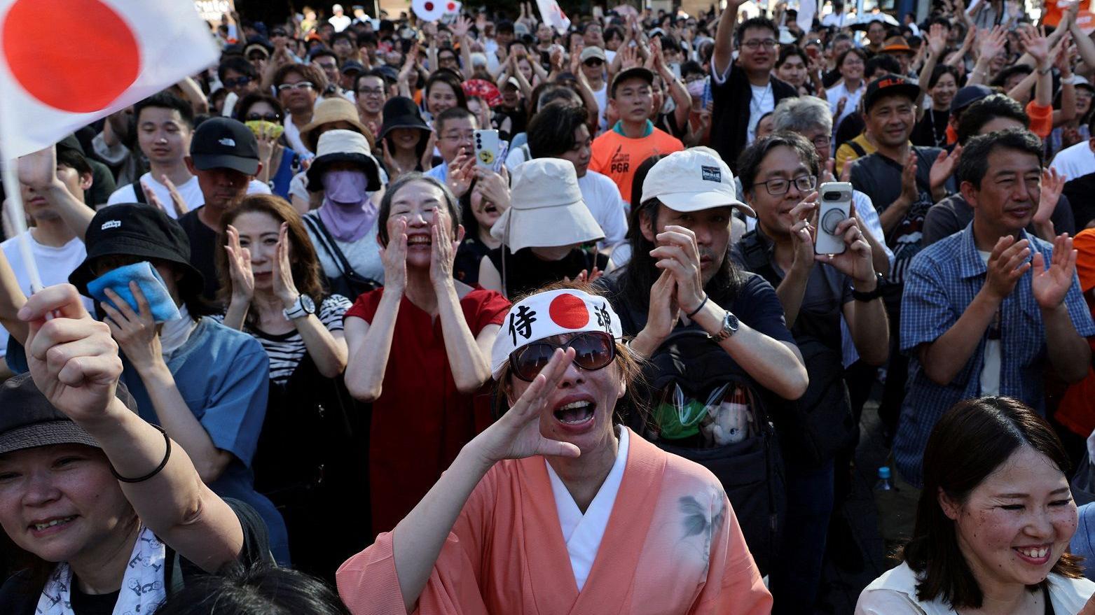 Pessoas vibram em evento ao ar livre, algumas com bandeiras s&iacute;mbolos do Jap&atilde;o; no centro, uma mulher com quimono tradicional 