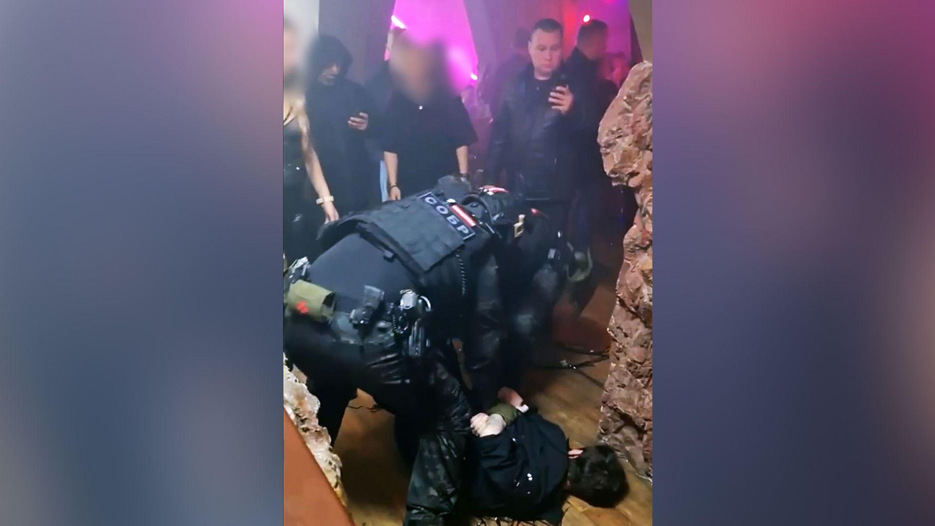 Um agente das for&ccedil;as de seguran&ccedil;a russas imobiliza uma pessoa no ch&atilde;o da festa, com os bra&ccedil;os para tr&aacute;s, enquanto outros convidados observam. A boate &eacute; iluminada por luzes de neon cor-de-rosa