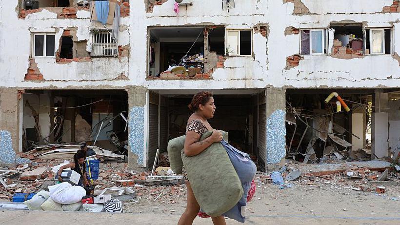 Una mujer carga enseres frente a un edificio con la fachada dañada por el terremoto. En segundo plano, otroas dos personas parecen cargar pertenencias. 
