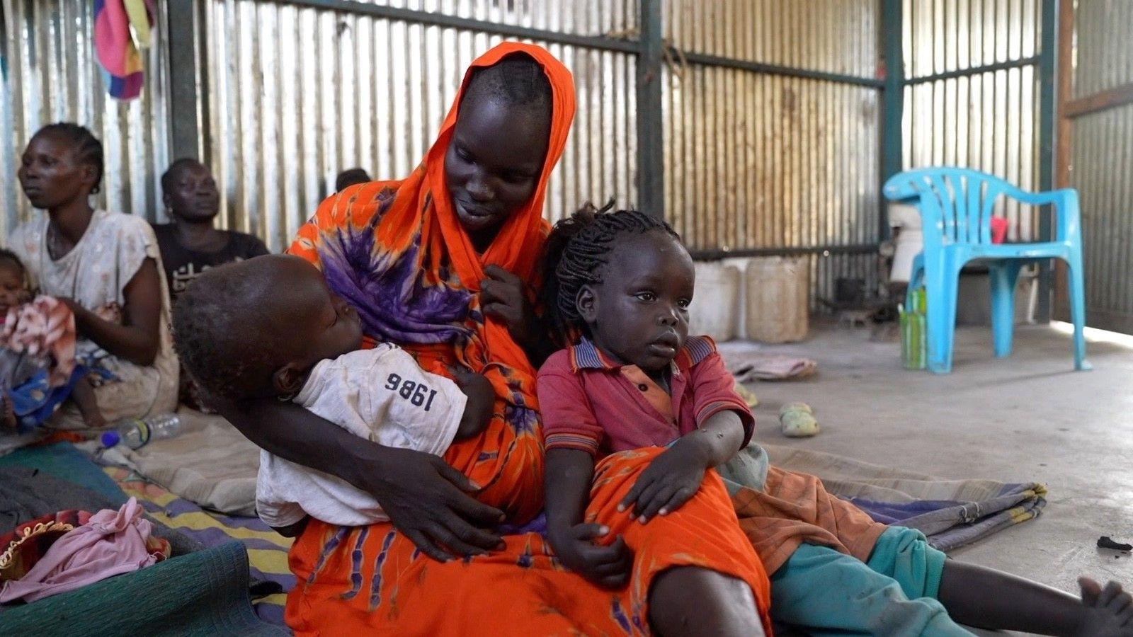 قصة أم سودانية عالقة هي وأطفالها بين صراعين 1 سارة ويليامز، مرتدية حجابًا برتقالي اللون، تجلس مع اثنين من أطفالها في حجرها
