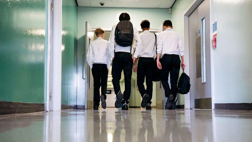 Cuatro chicos de escuela secundaria, con camisas blancas y pantalones negros, se ven de espalda cargando sus mochilas y dirigiéndose a clase dentro del edificio escolar, uno al lado del otro