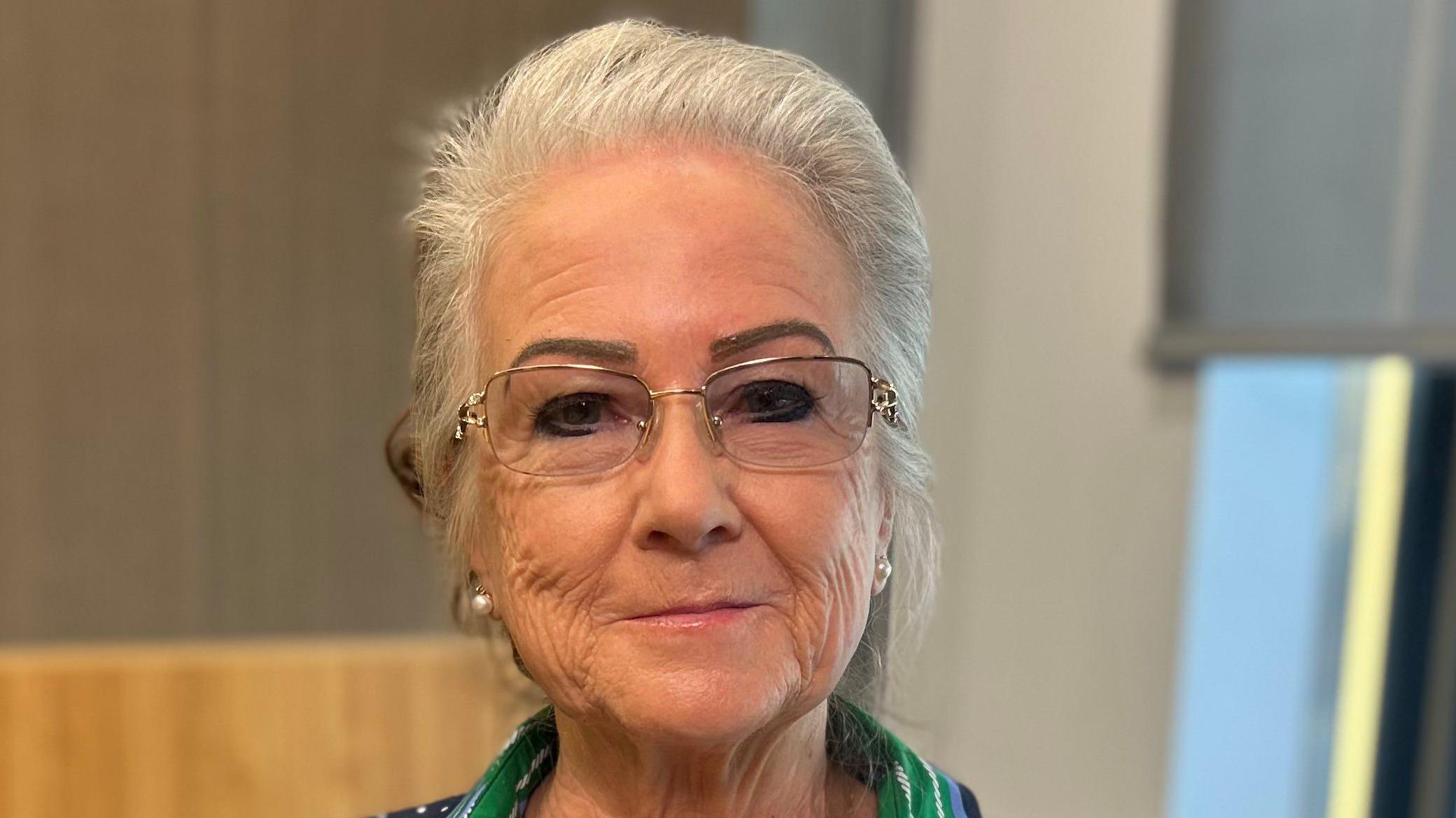 Uma mulher de cabelos prateados olha para a câmera. Ela usa óculos pequenos com armação prateada.