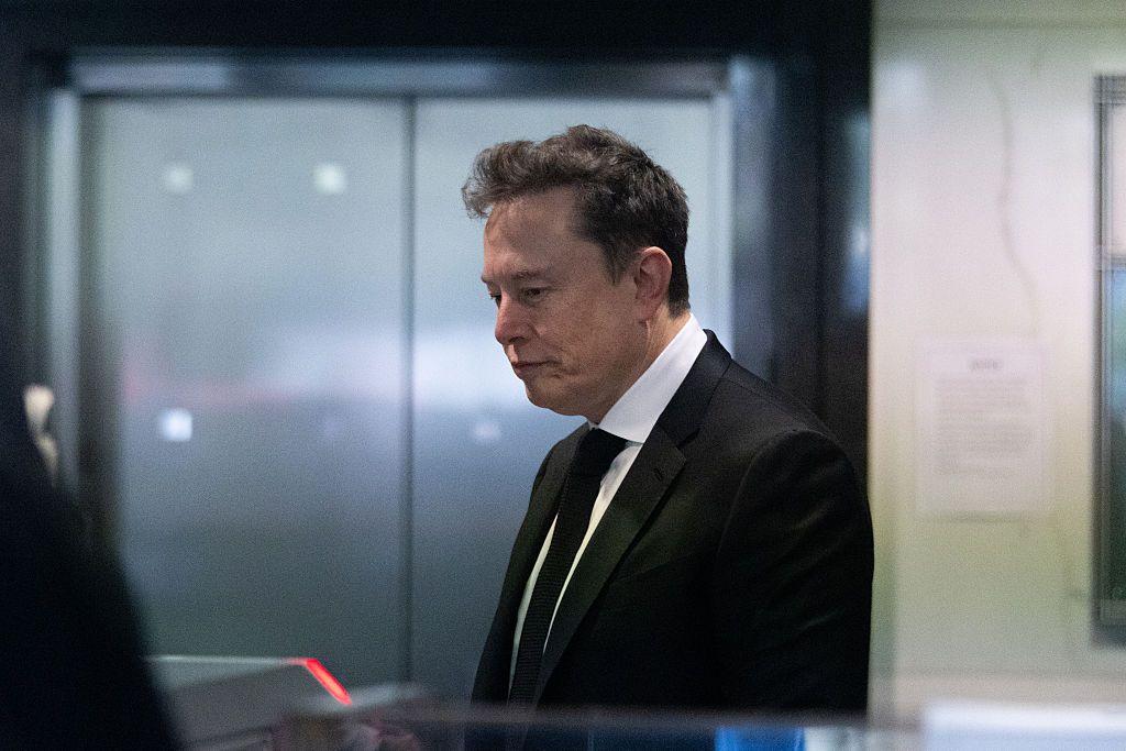 Elon Musk con traje negro y corbata, de perfil, en un interior moderno con paredes de vidrio.