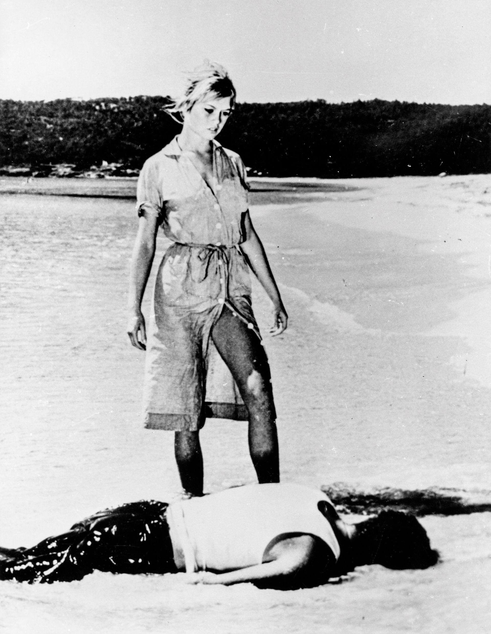 Brigitte Bardot em uma cena do filme E Deus Criou a Mulher, usando um vestido parcialmente desabotoado na barra, que se abre para revelar sua coxa, enquanto ela est&aacute; em p&eacute; sobre um homem deitado de bru&ccedil;os na praia.