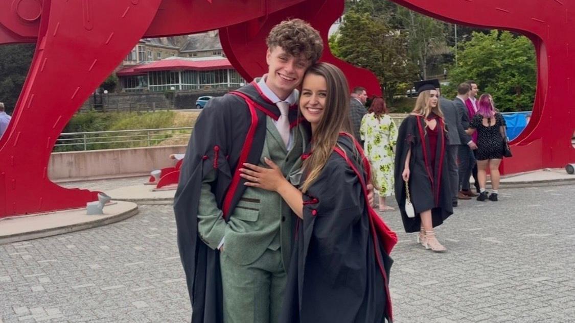 Hannah e Jack est&atilde;o sorrindo para a c&acirc;mera, lado a lado. Ambos vestem becas universit&aacute;rias vermelhas e pretas. Jack tamb&eacute;m usa um terno verde claro.