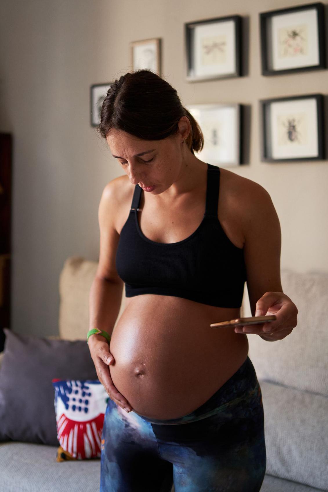 Una mujer embarazada está de pie y toca su barriga  mientras sostiene con la otra mano un celular
