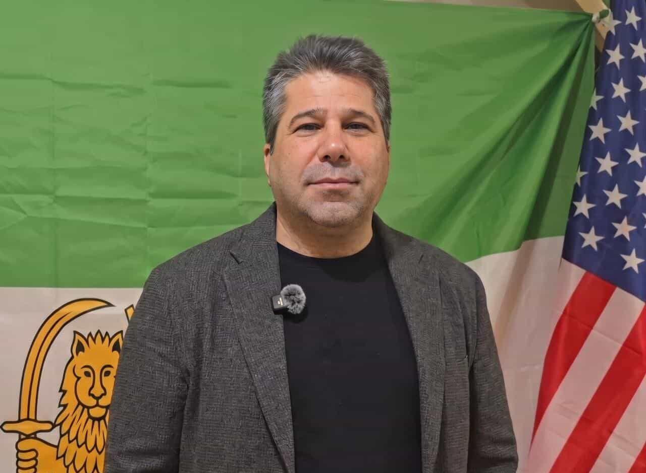  مسعود طاہری کی تصویر، پس منظر میں شیر اور سورجوالا پرچم اور امریکی پرچم دکھائی دے رہے ہیں