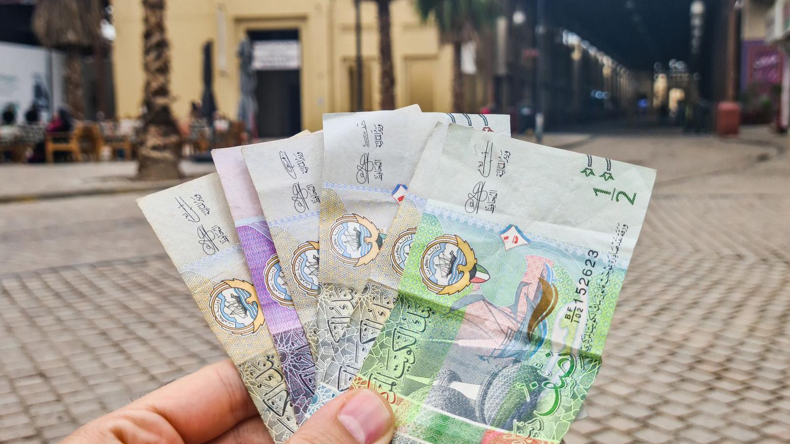 صورة ليد رجل تحمل دنانير كويتية ويبرز في خلفية الصورة شارع.