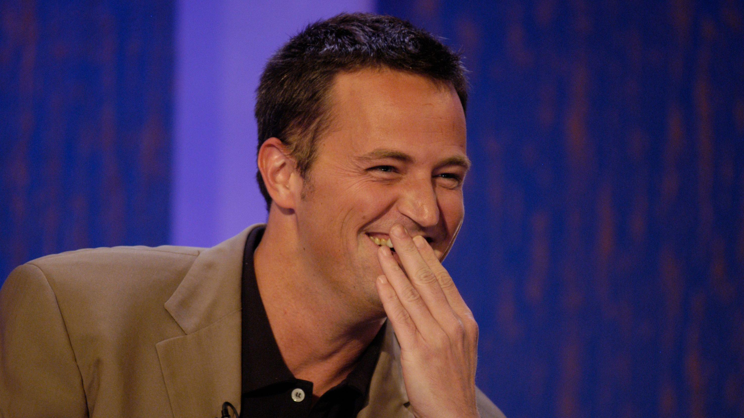 Matthew Perry cobre a boca enquanto ri. Ele veste um blazer bege e uma camiseta polo preta.