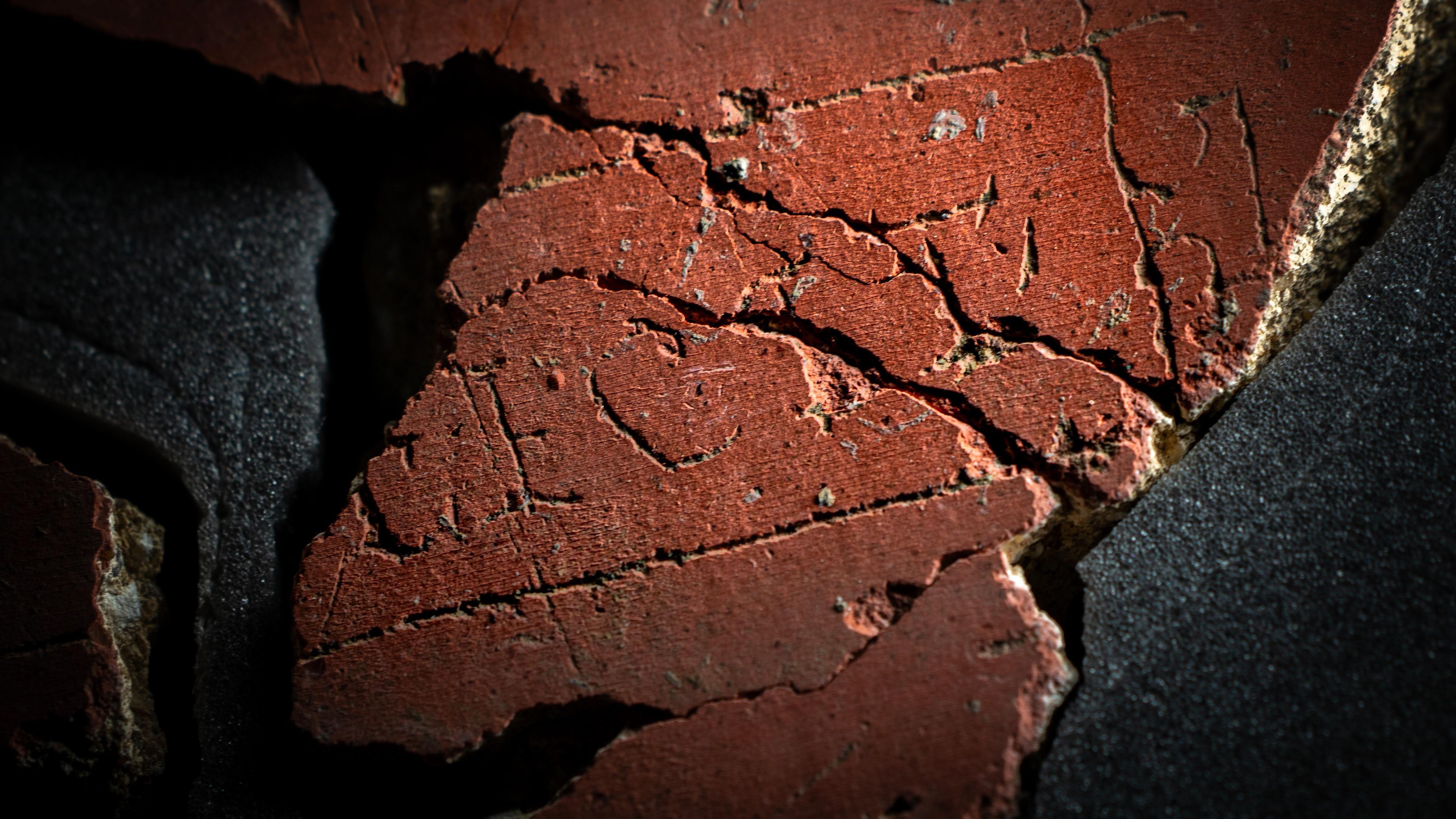 Un primer plano de un trozo de yeso rojo, formado por varios trozos más pequeños, con las letras F E C I T en mayúsculas claramente visibles con una línea dibujada en forma de cuadrado alrededor de tres lados de la palabra. Falta el lado izquierdo del yeso, donde debería estar el nombre de la persona o grupo que realizó la obra de arte.