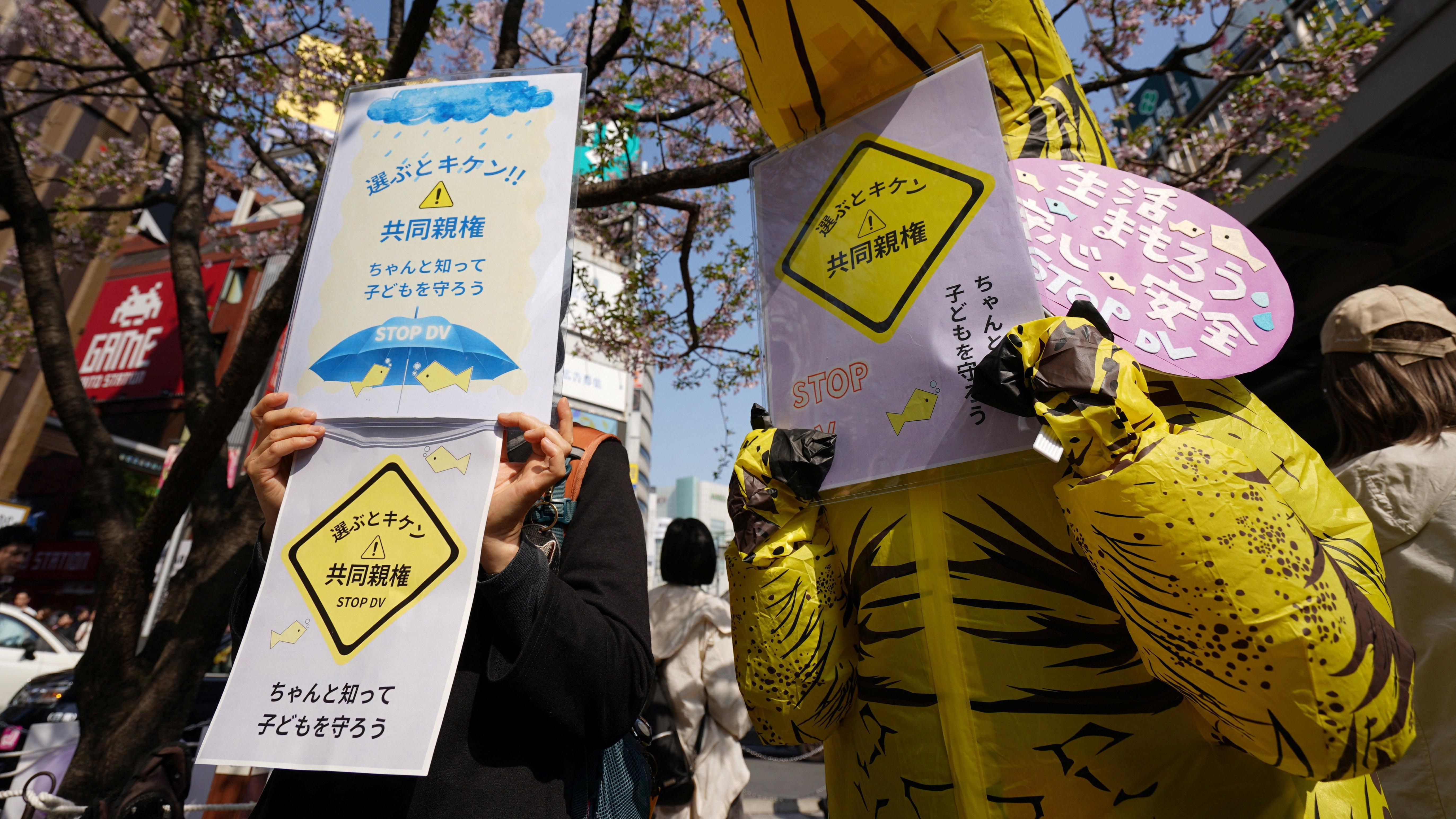 日本反家暴團體成員在東京新宿戰士反對《民法》修正案「共同親權」條款標語牌，其中一人（右）身穿恐龍裝束（29/3/2026）