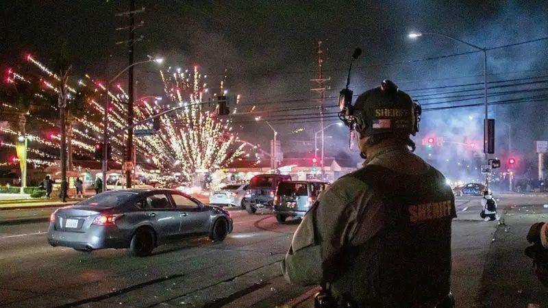 Um agente parado olhando para carros passando. Ao fundo há fogos de artifício